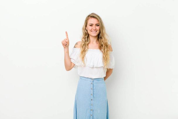 young blonde woman smiling cheerfully and happily, pointing upwards with one hand to copy space against white wall - Fotografie, Obrázek