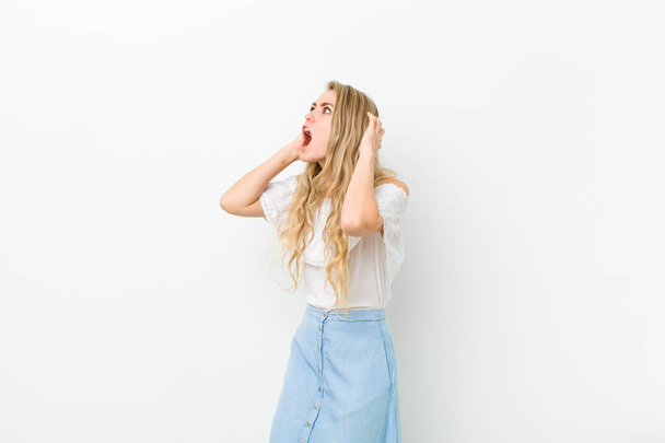 young blonde woman with open mouth, looking horrified and shocked because of a terrible mistake, raising hands to head against white wall - Fotoğraf, Görsel