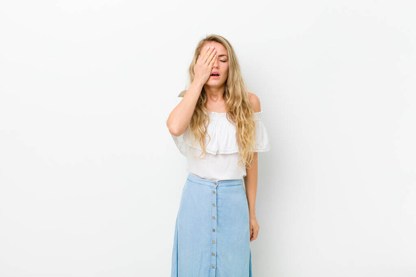 young blonde woman looking sleepy, bored and yawning, with a headache and one hand covering half the face against white wall - Фото, изображение