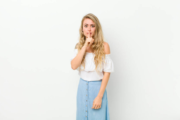 young blonde woman asking for silence and quiet, gesturing with finger in front of mouth, saying shh or keeping a secret against white wall - Fotoğraf, Görsel