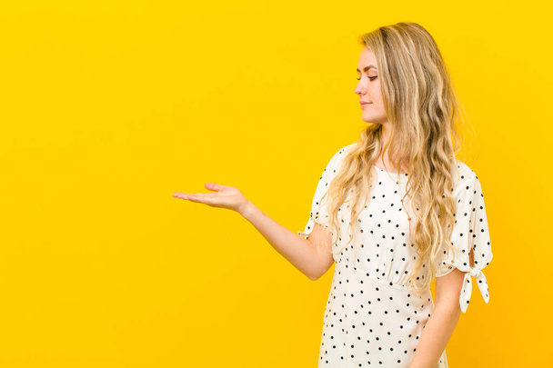 young blonde woman feeling happy and smiling casually, looking to an object or concept held on the hand on the side against yellow wall - 写真・画像