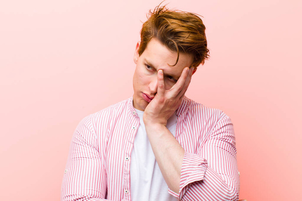 young red head man feeling bored, frustrated and sleepy after a tiresome, dull and tedious task, holding face with hand against pink wall - Photo, image