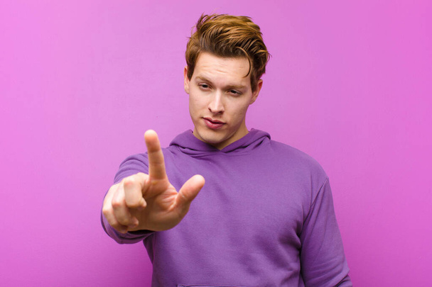 young red head man smiling proudly and confidently making number one pose triumphantly, feeling like a leader against purple wall - Фото, изображение