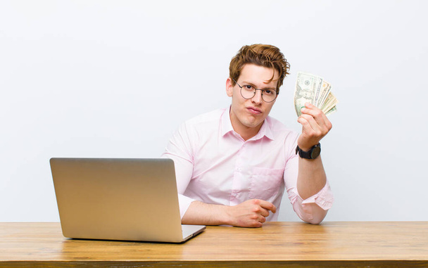 junger Geschäftsmann mit rotem Kopf, der in seinem Schreibtisch arbeitet, Geldkonzept - Foto, Bild