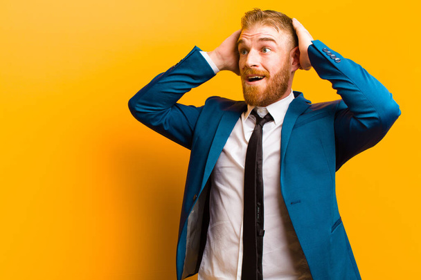 young red head businessman with open mouth, looking horrified and shocked because of a terrible mistake, raising hands to head against orange background - Photo, Image