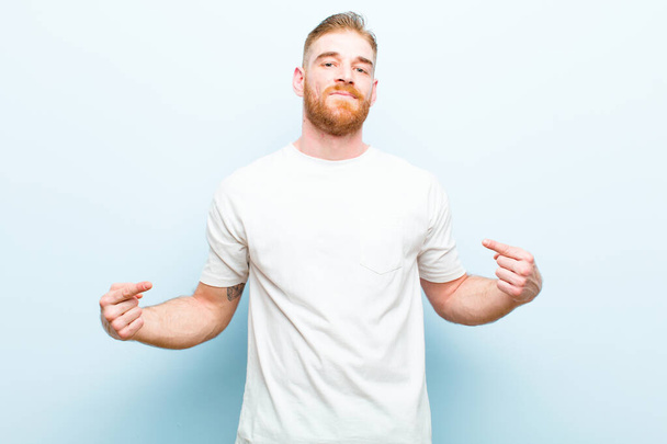 young red head man looking proud, arrogant, happy, surprised and satisfied, pointing to self, feeling like a winner against soft blue background - 写真・画像