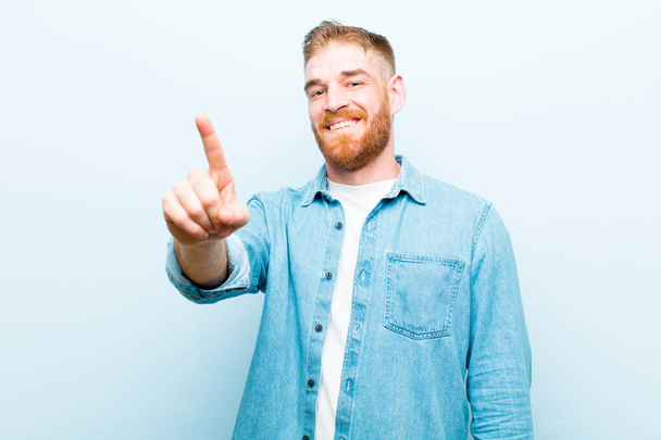 young red head man smiling proudly and confidently making number one pose triumphantly, feeling like a leader against soft blue background - Φωτογραφία, εικόνα