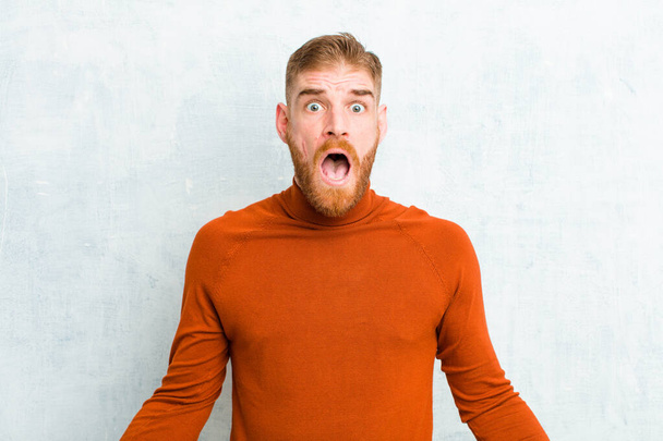 young red head man wearing turtle neck feeling terrified and shocked, with mouth wide open in surprise against cement wall - Фото, изображение
