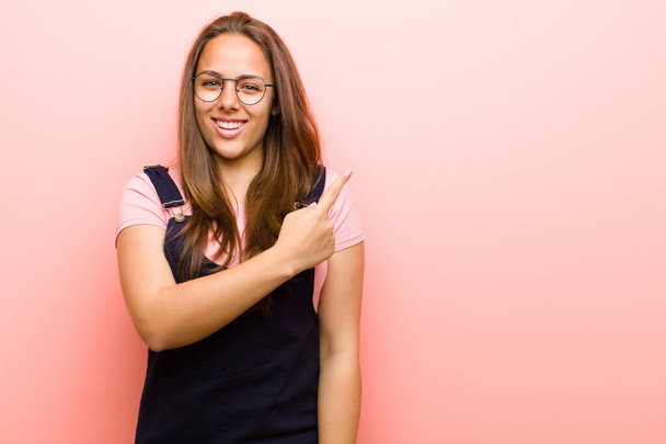 jonge vrouw glimlachen vrolijk, zich gelukkig voelen en wijzen naar de zijkant en naar boven, tonen object in kopieerruimte tegen roze achtergrond - Foto, afbeelding