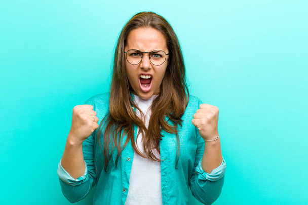 young woman shouting aggressively with annoyed, frustrated, angry look and tight fists, feeling furious against blue background - Foto, Imagen