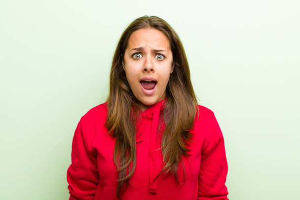 young woman feeling terrified and shocked, with mouth wide open in surprise against green background - Foto, immagini