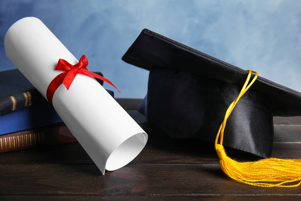Graduation hat, books and student's diploma on black wooden table against light blue background - Fotó, kép