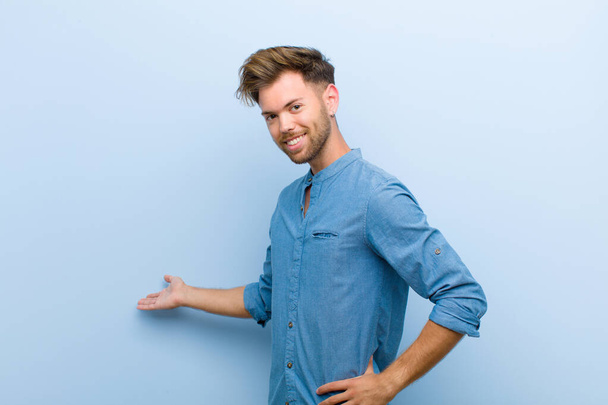 young businessman feeling happy and cheerful, smiling and welcoming you, inviting you in with a friendly gesture against blue background - Φωτογραφία, εικόνα