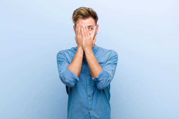 young businessman covering face with hands, peeking between fingers with surprised expression and looking to the side against blue background - 写真・画像
