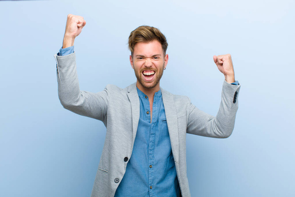 young businessman shouting triumphantly, looking like excited, happy and surprised winner, celebrating against blue background - Photo, Image