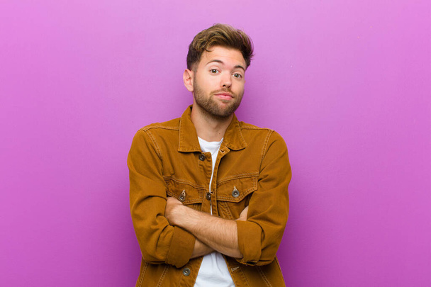 young man shrugging, feeling confused and uncertain, doubting with arms crossed and puzzled look against purple background - Photo, image