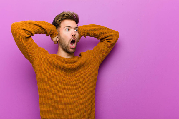 young man with open mouth, looking horrified and shocked because of a terrible mistake, raising hands to head against purple background - Fotografie, Obrázek