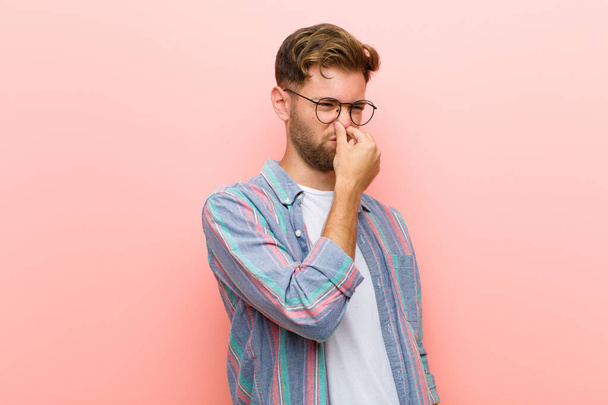 young man feeling disgusted, holding nose to avoid smelling a foul and unpleasant stench against pink background - 写真・画像