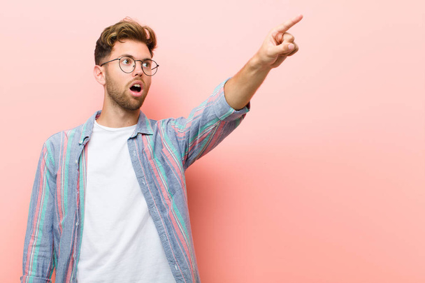 young man feeling shocked and surprised, pointing and looking upwards in awe with amazed, open-mouthed look against pink background - Foto, imagen
