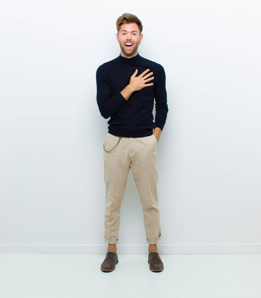 young full body man feeling shocked and surprised, smiling, taking hand to heart, happy to be the one or showing gratitude against white background - Foto, imagen