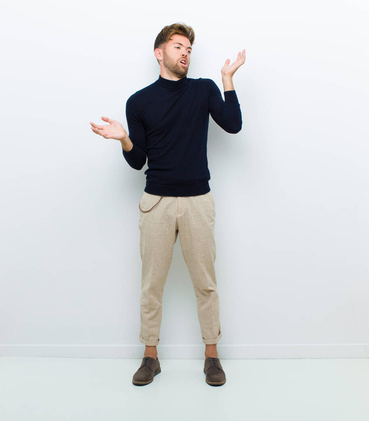 young full body man shrugging with a dumb, crazy, confused, puzzled expression, feeling annoyed and clueless against white background - Photo, Image