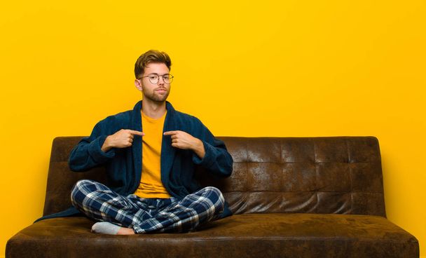 young man wearing pajamas looking proud, positive and casual pointing to chest with both hands . sitting on a sofa - Photo, Image