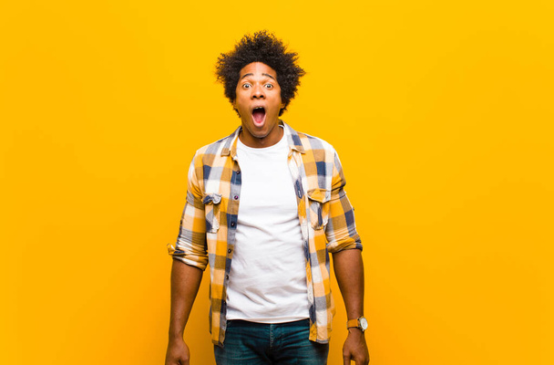 young black man looking shocked, angry, annoyed or disappointed, open mouthed and furious against orange wall - Fotoğraf, Görsel