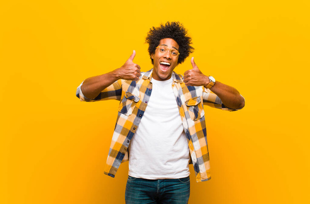 young black man smiling broadly looking happy, positive, confident and successful, with both thumbs up against orange wall - Foto, imagen