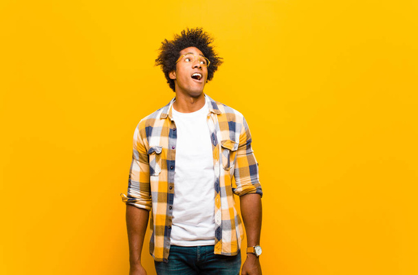 young black man feeling shocked, happy, amazed and surprised, looking to the side with open mouth against orange wall - Fotoğraf, Görsel