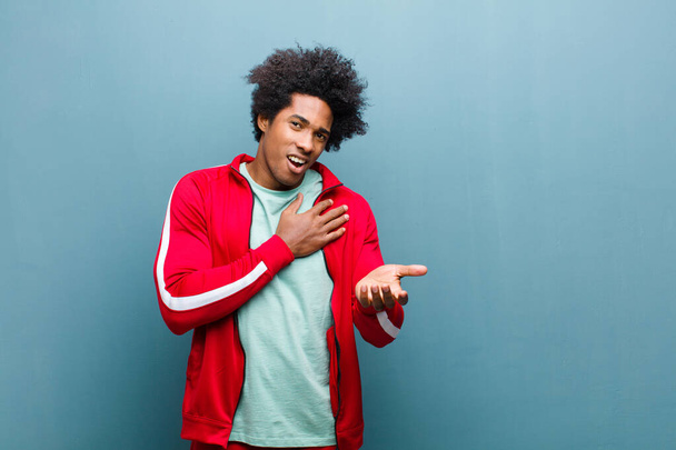 young black sports man feeling happy and in love, smiling with one hand next to heart and the other stretched up front against grunge wall - Photo, Image