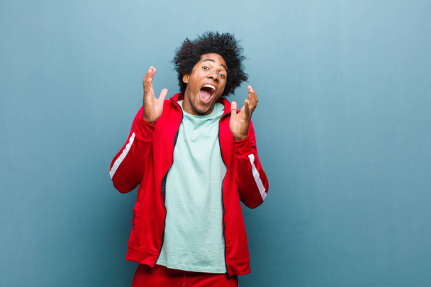 young black sports man feeling shocked and excited, laughing, amazed and happy because of an unexpected surprise against grunge wall - Fotoğraf, Görsel