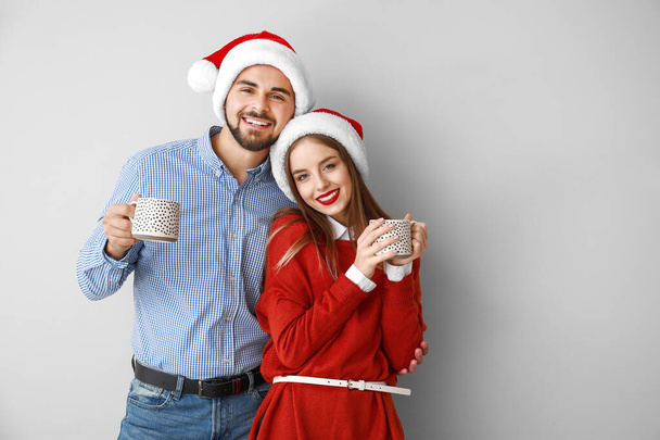 Happy young couple in Santa hats and with hot chocolate on light background - Φωτογραφία, εικόνα