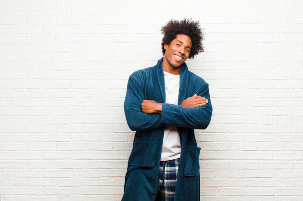 young black man wearing pajamas with gown looking like a happy, proud and satisfied achiever smiling with arms crossed against brick wall - Valokuva, kuva