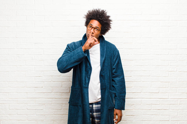 young black man wearing pajamas with gown asking for silence and quiet, gesturing with finger in front of mouth, saying shh or keeping a secret against brick wall - Foto, afbeelding