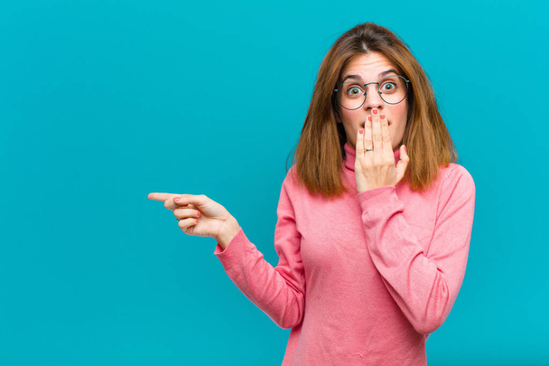 young pretty woman feeling happy, shocked and surprised, covering mouth with hand and pointing to lateral copy space against blue background - Foto, Bild