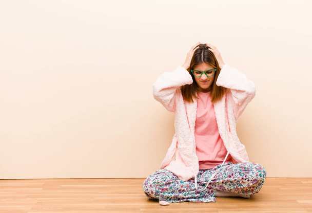 young woman wearing pajamas sitting at home feeling stressed and frustrated, raising hands to head, feeling tired, unhappy and with migraine - Fotografie, Obrázek