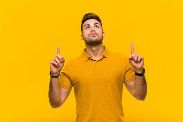 young hispanic man looking shocked, amazed and open mouthed, pointing upwards with both hands to copy space against orange wall - Foto, imagen