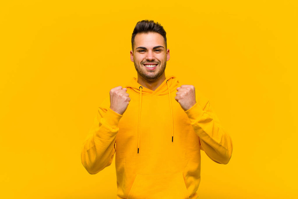 young hispanic man shouting triumphantly, laughing and feeling happy and excited while celebrating success against orange wall - Фото, изображение