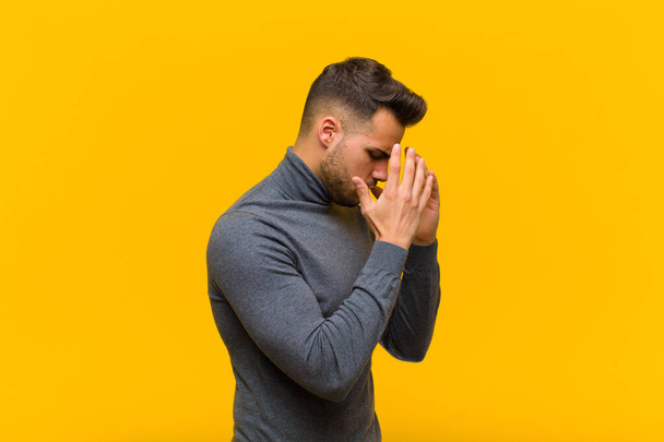 young hispanic man covering eyes with hands with a sad, frustrated look of despair, crying, side view against orange wall - Fotoğraf, Görsel