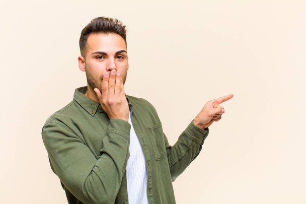 young hispanic man feeling happy, shocked and surprised, covering mouth with hand and pointing to lateral copy space against isolated background - Fotoğraf, Görsel