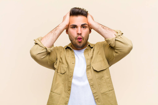 young hispanic man looking excited and surprised, open-mouthed with both hands on head, feeling like a lucky winner against isolated background - Foto, Imagen
