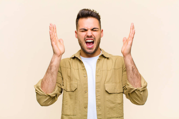 young hispanic man furiously screaming, feeling stressed and annoyed with hands up in the air saying why me against isolated background - Foto, imagen