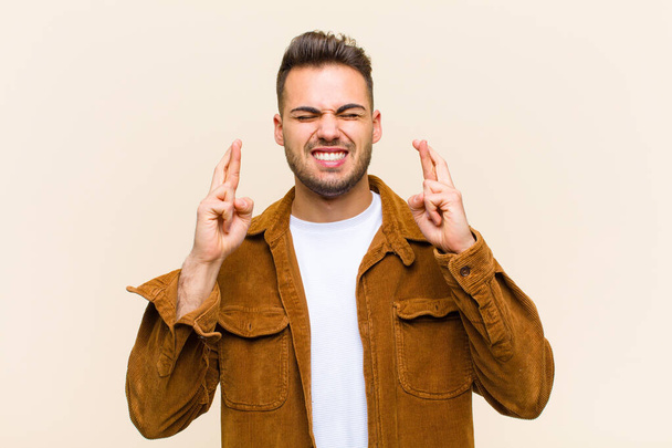 young hispanic man smiling and anxiously crossing both fingers, feeling worried and wishing or hoping for good luck against isolated background - Фото, зображення
