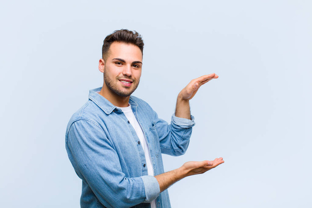 young hispanic man smiling, feeling happy, positive and satisfied, holding or showing object or concept on copy space against blue wall - Φωτογραφία, εικόνα