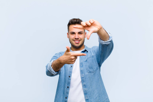 young hispanic man feeling happy, friendly and positive, smiling and making a portrait or photo frame with hands against blue wall - 写真・画像