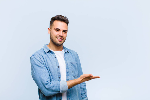 young hispanic man smiling cheerfully, feeling happy and showing a concept in copy space with palm of hand against blue wall - Fotó, kép