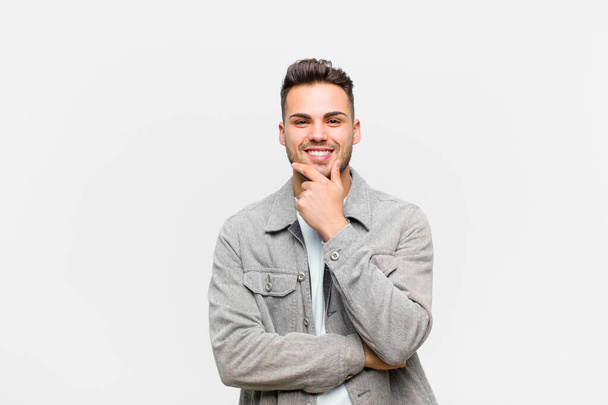 young hispanic man smiling, enjoying life, feeling happy, friendly, satisfied and carefree with hand on chin against gray background - Φωτογραφία, εικόνα