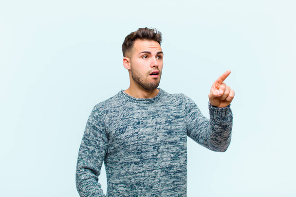 young hispanic man feeling shocked and surprised, pointing and looking upwards in awe with amazed, open-mouthed look against blue background - Photo, image