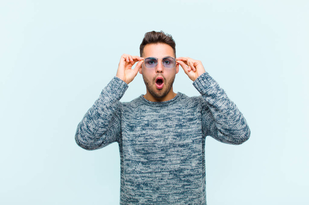 young hispanic man feeling shocked, amazed and surprised, holding glasses with astonished, disbelieving look against blue background - Foto, imagen
