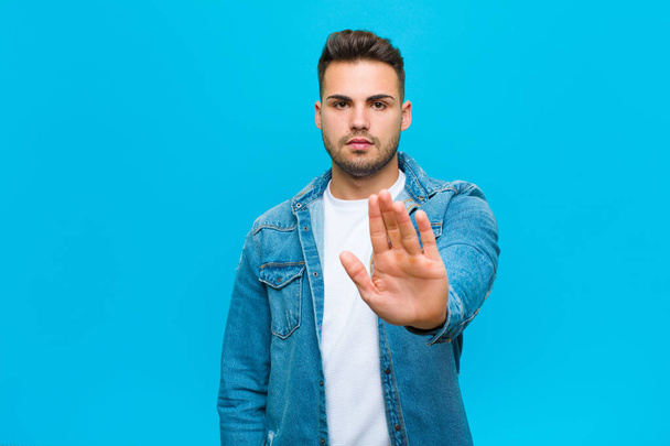 young hispanic man looking serious, stern, displeased and angry showing open palm making stop gesture against blue background - Фото, зображення